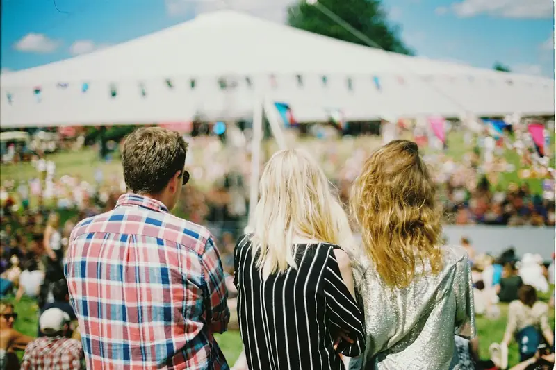 Three friends in a festival crowd facing a white marquee hung with bunting.