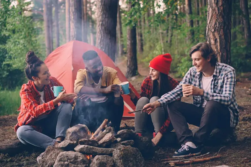 Four friends drinking from mugs around a campfire beside a tent in a pine wood.